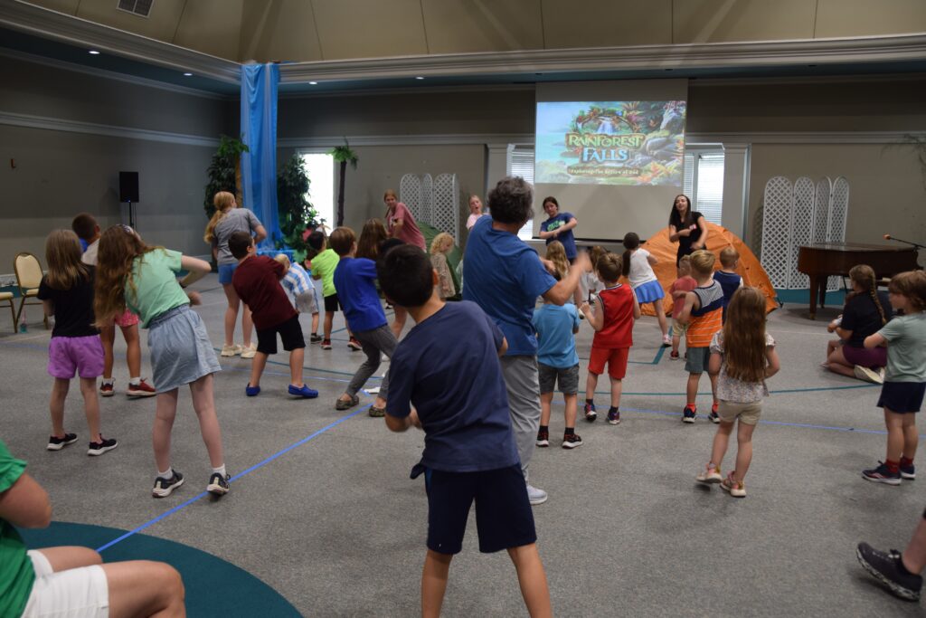 Group of children dancing in a large hall with a rainforest-themed slide projected on a screen at the front.