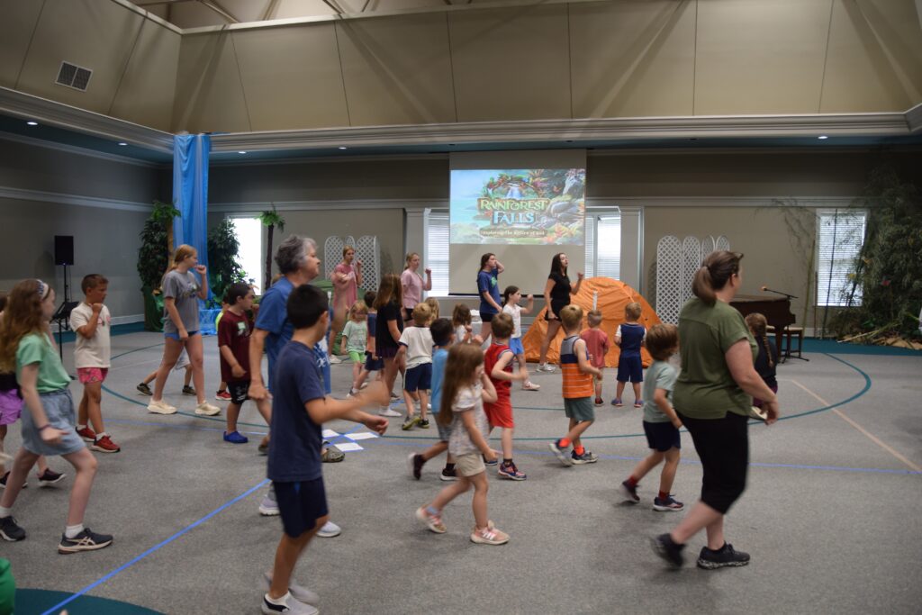 Children walking and standing around in a large indoor hall with a tropical stage setup and a projector showing a rainforest-themed image in the background.