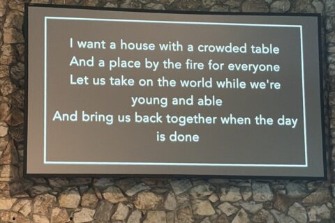 Screen showing song lyrics projected on a stone wall, including 'I want a house with a crowded table' lines, with an exit sign underneath.