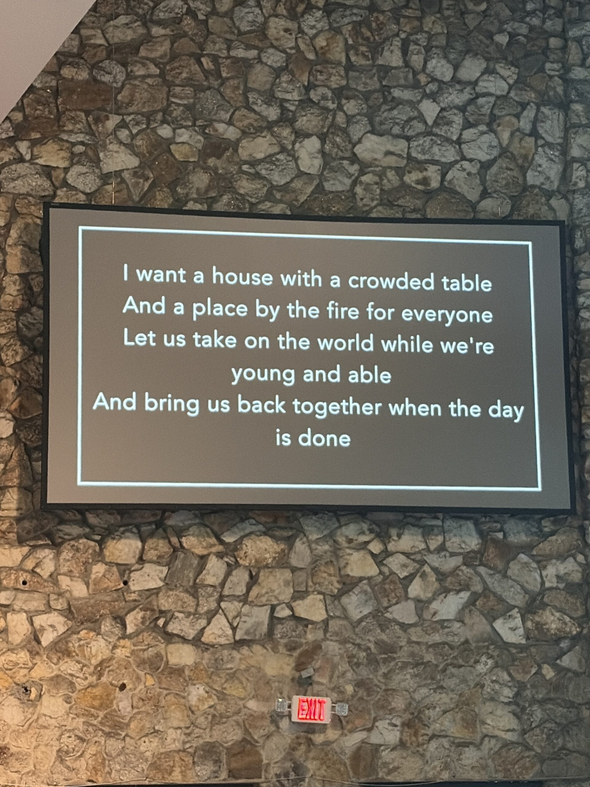 Screen showing song lyrics projected on a stone wall, including 'I want a house with a crowded table' lines, with an exit sign underneath.