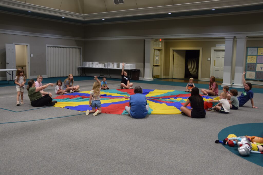 Children and adults sitting in a circle on a large rainbow-colored parachute indoors, likely playing a group game in a community space.