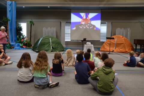 Children sit cross‑legged on a gym floor watching a projector screen showing two people on a couch, with green and orange tents set up for a camp-themed event in the background.