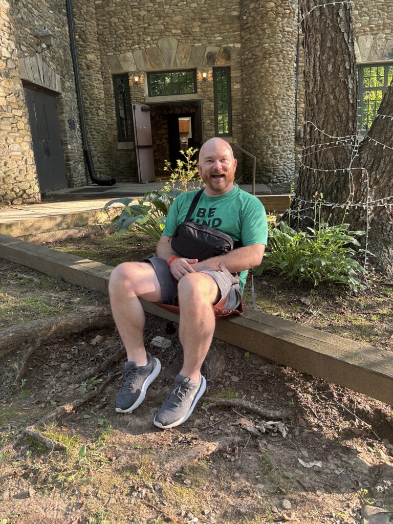 Smiling man in a green t-shirt with a crossbody bag sits on a wooden beam outside a stone building entrance.