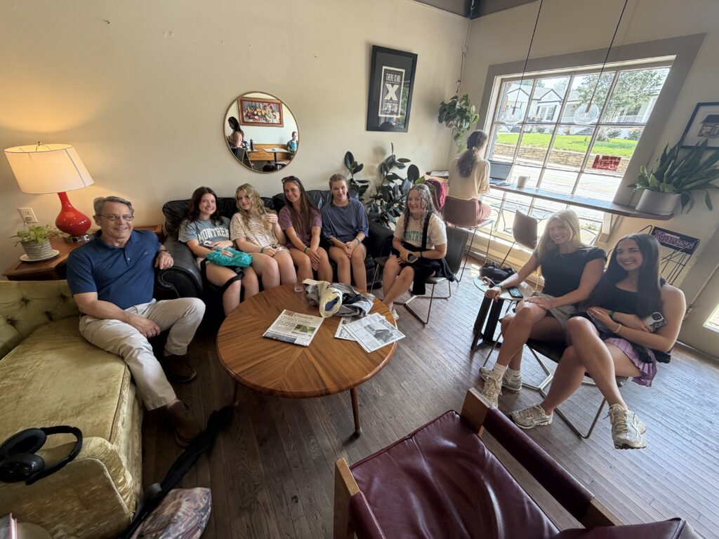Group of people relaxing in a bright cafe lounge, seated around a round coffee table with newspapers nearby.