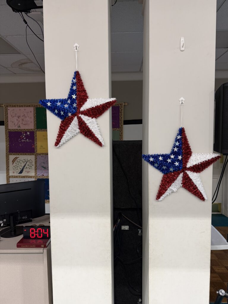 Patriotic red, white, and blue star garlands dangling from white columns in a room, with an electronic clock and classroom boards in the background.