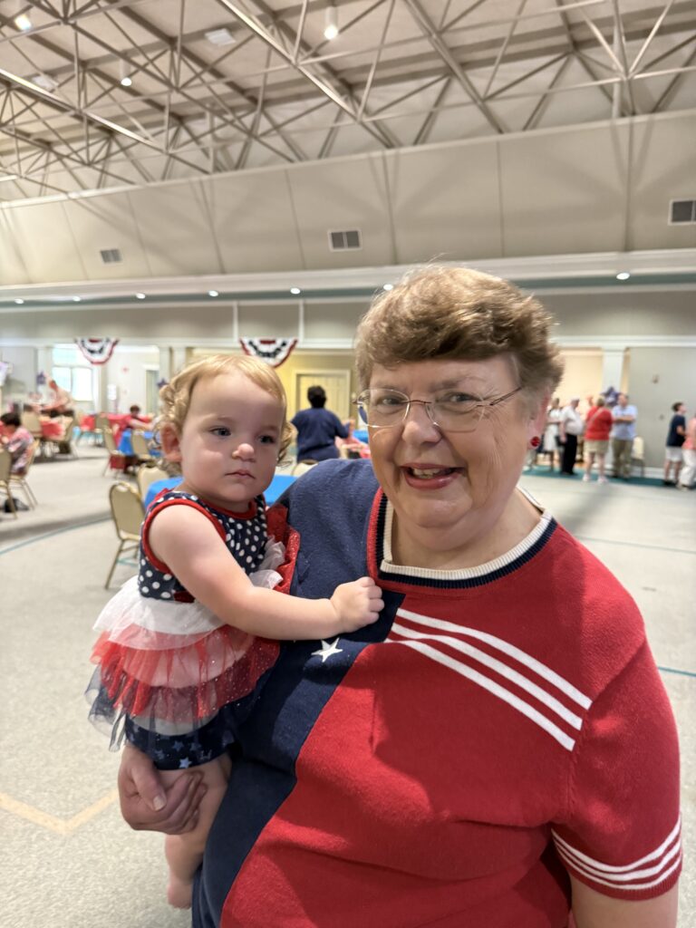 Smiling older woman in a red, white, and blue top holding a toddler in a patriotic dress at an indoor event.