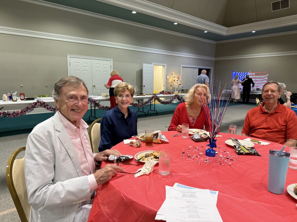 Group of seniors seated around a round table with a red tablecloth, eating and chatting at a holiday party in a banquet hall.