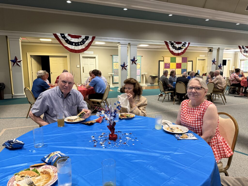 People seated at tables with blue tablecloths at a patriotic community event, red, white, and blue bunting overhead.