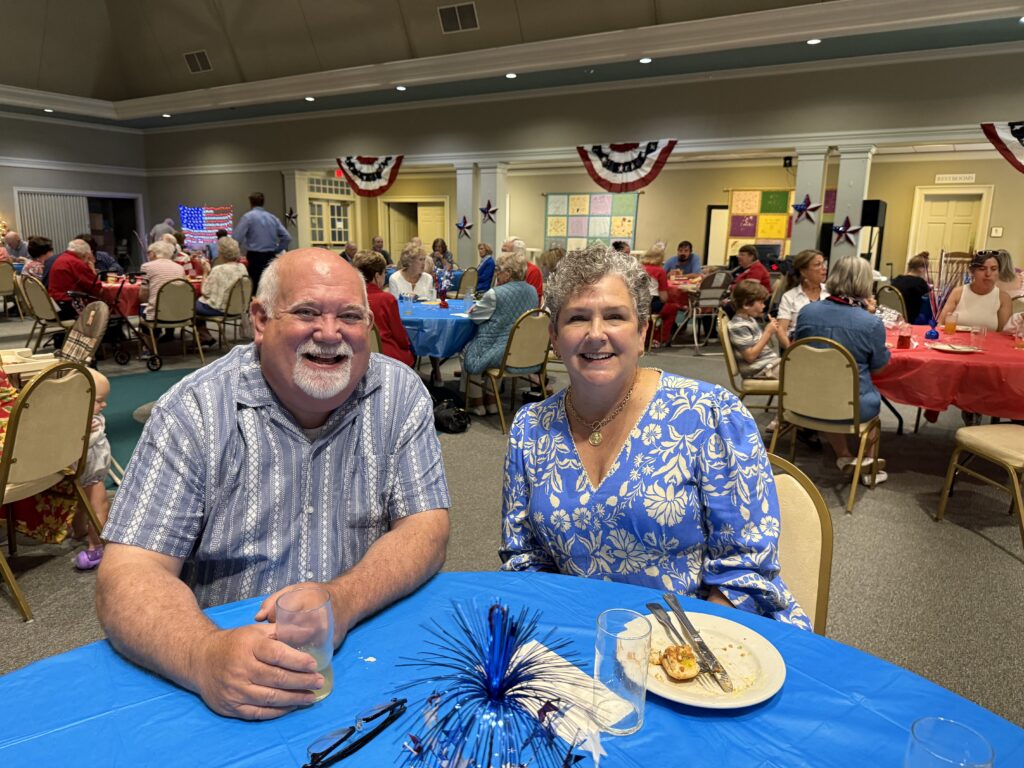 Smiling couple at a blue-tablecloth table at a large community event hall with red, white, and blue bunting and many guests in the background.