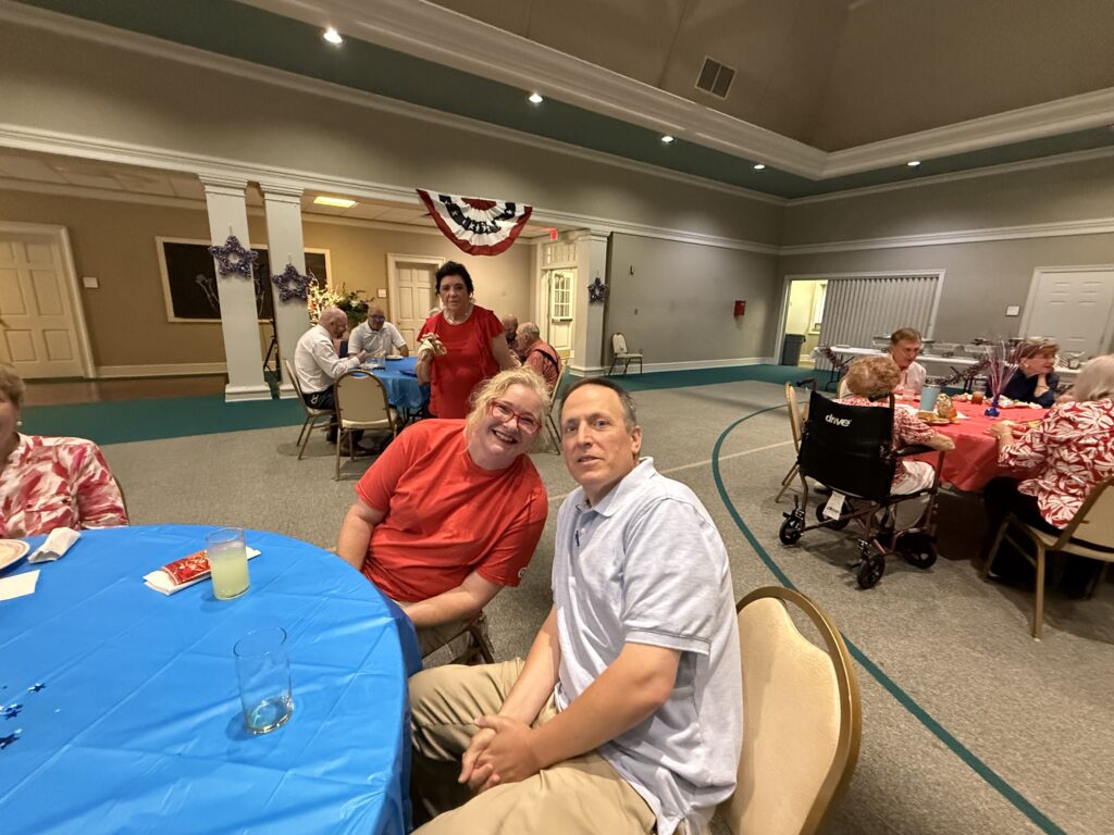 Group of seniors at a festive indoor event, seated around blue tables with red dress attire and snacks visible.