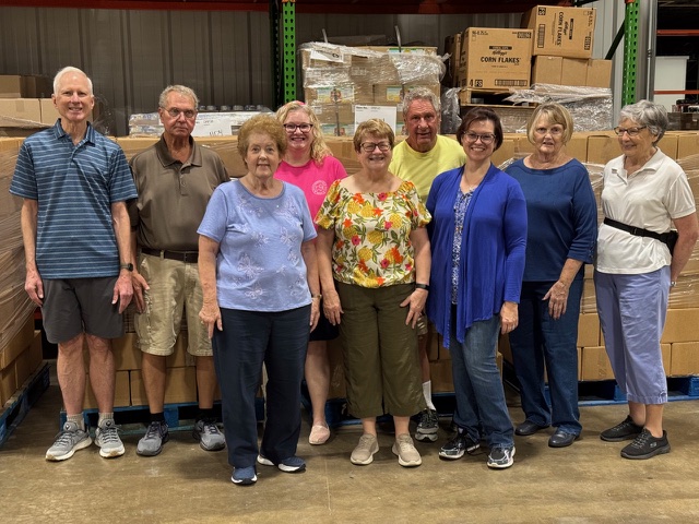 Group of ten adults standing in a warehouse with stacked boxes and pallets in the background, posing for a photo.