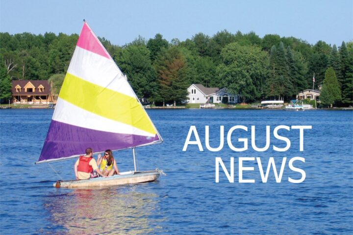 Colorful sailboat with two people in life jackets on a blue lake, background houses and trees, large white text reading AUGUST NEWS overlaying the water.